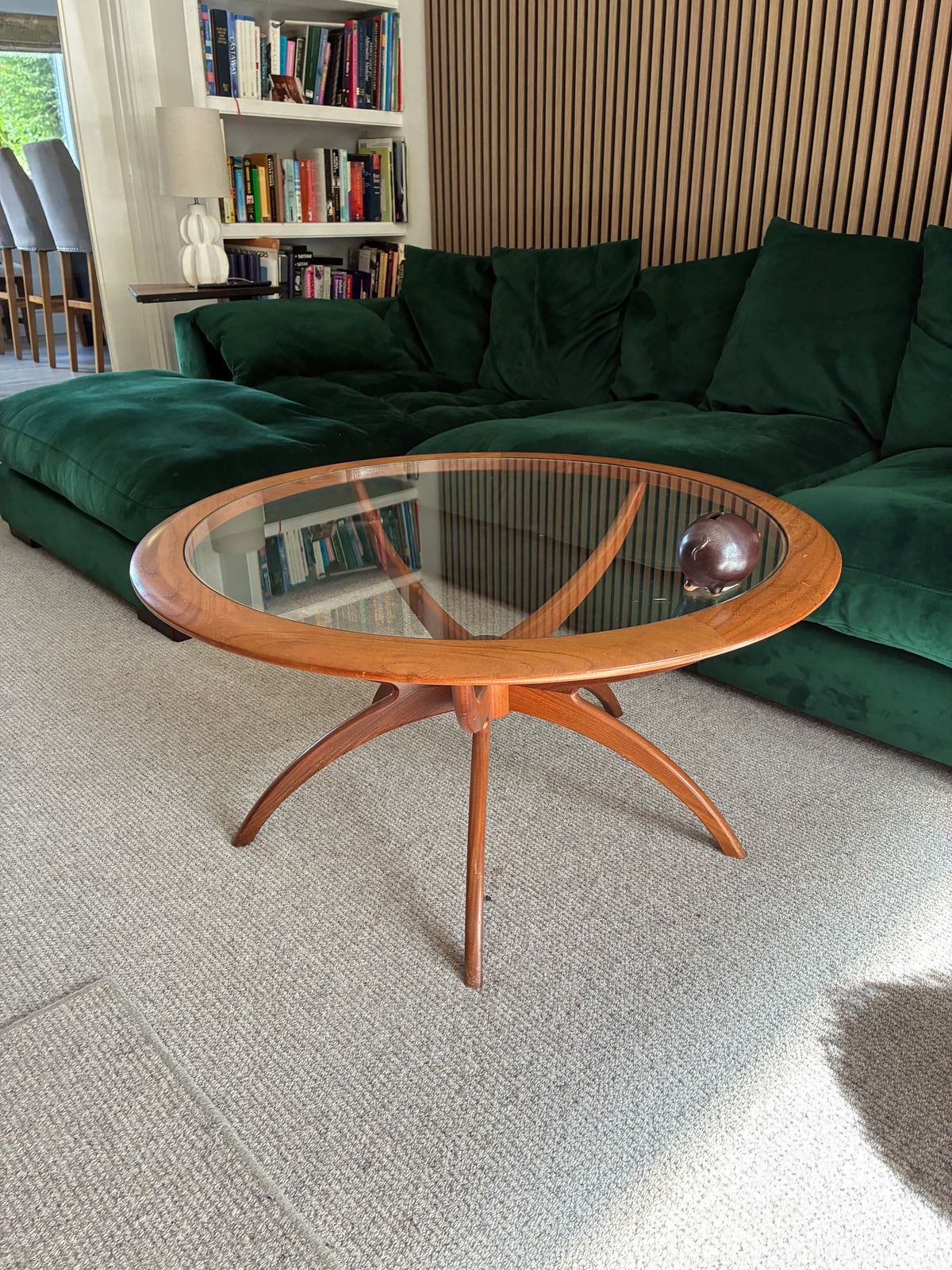 A mid-century modern g plan spider wooden coffee table with a clear glass top and curved star-shaped legs, positioned in a living room in front of a green velvet sofa.