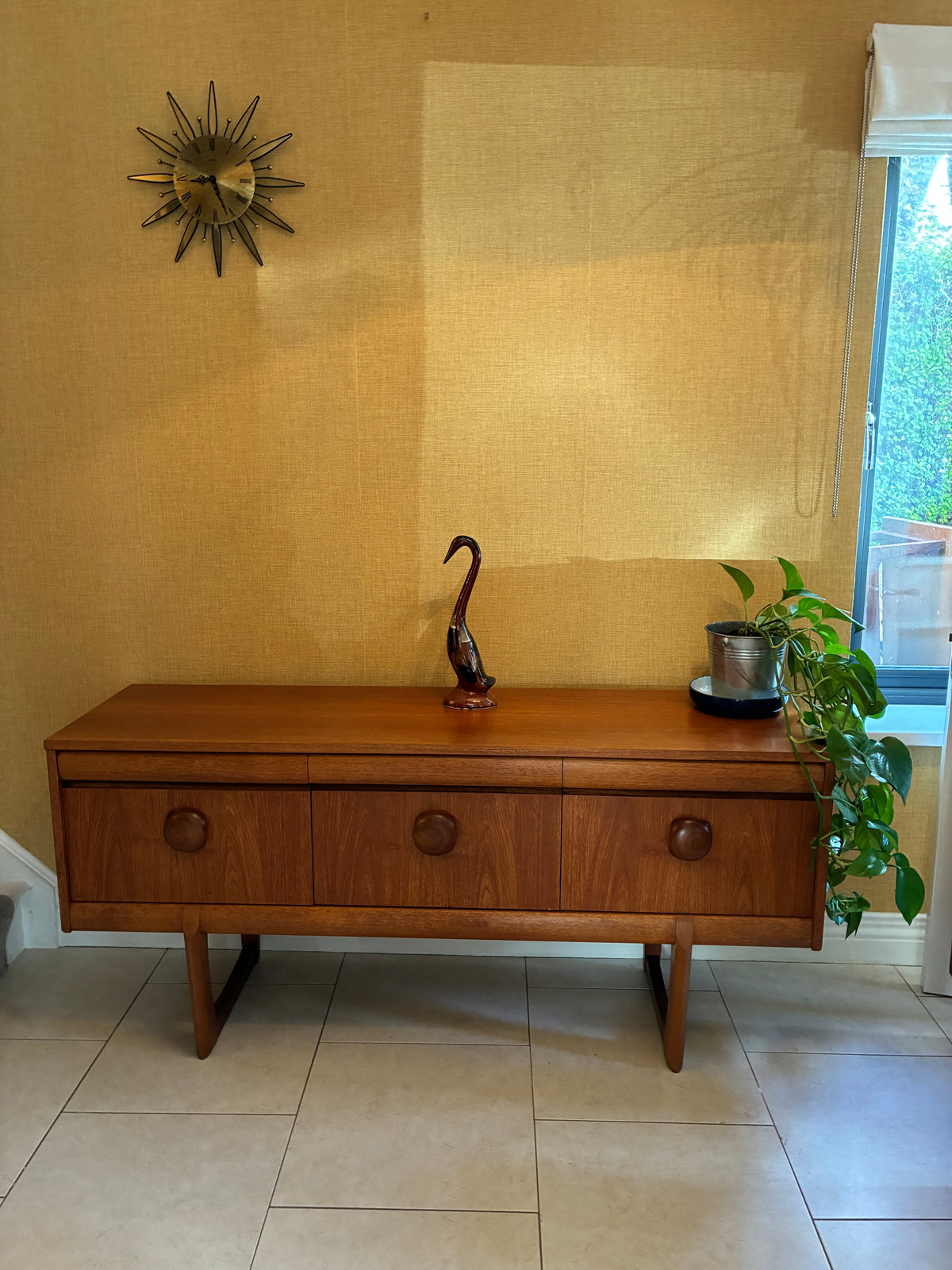 A mid-century modern wooden sideboard with three drawers and round knobs, styled in a home entryway against a textured yellow wall with a sunburst mirror and a potted plant.