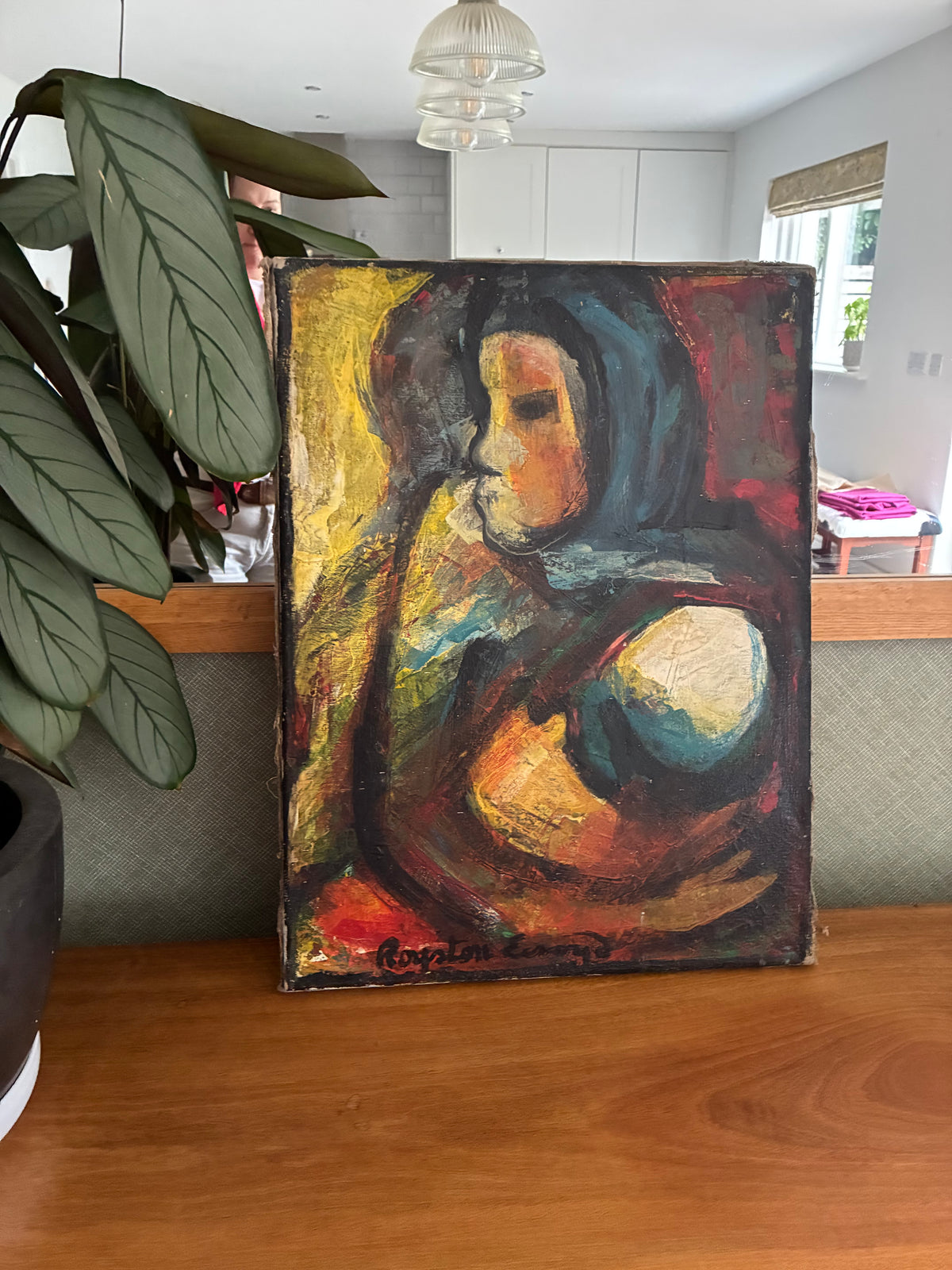 An expressive, colorful painting of a mother and child, displayed on a wooden table in a home interior next to a large green plant.