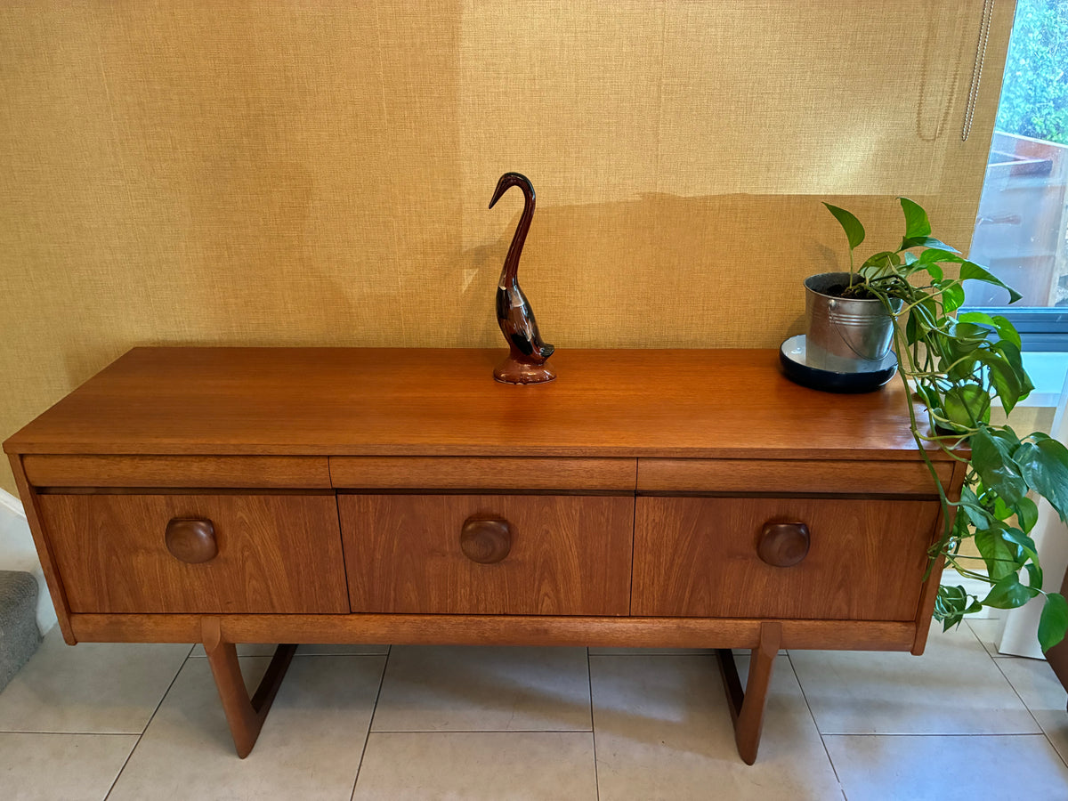 A mid-century modern wooden sideboard with three drawers and tapered legs, styled with a dark wood swan figurine and a potted green plant against a beige textured wall.