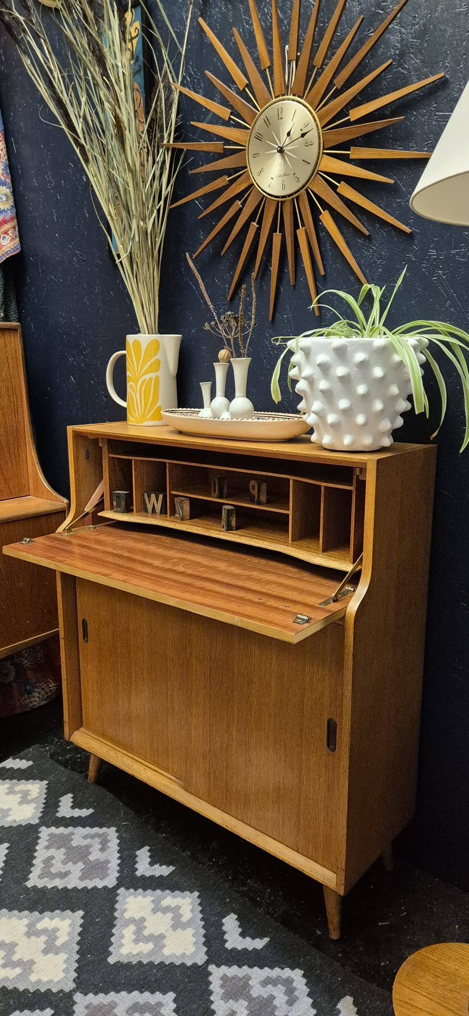 A mid-century modern wooden roll-top secretary cabinet with its door open, styled with decorative items including a potted plant, a textured vase, and a yellow mug on top, set against a dark blue wall with a sunburst clock.