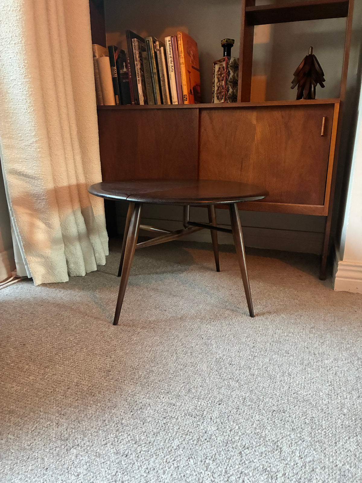 A mid-century modern ercol wooden side table with a round dark top and angled legs, positioned in a room with a beige carpet, a curtain, and a mid-century credenza in the background.