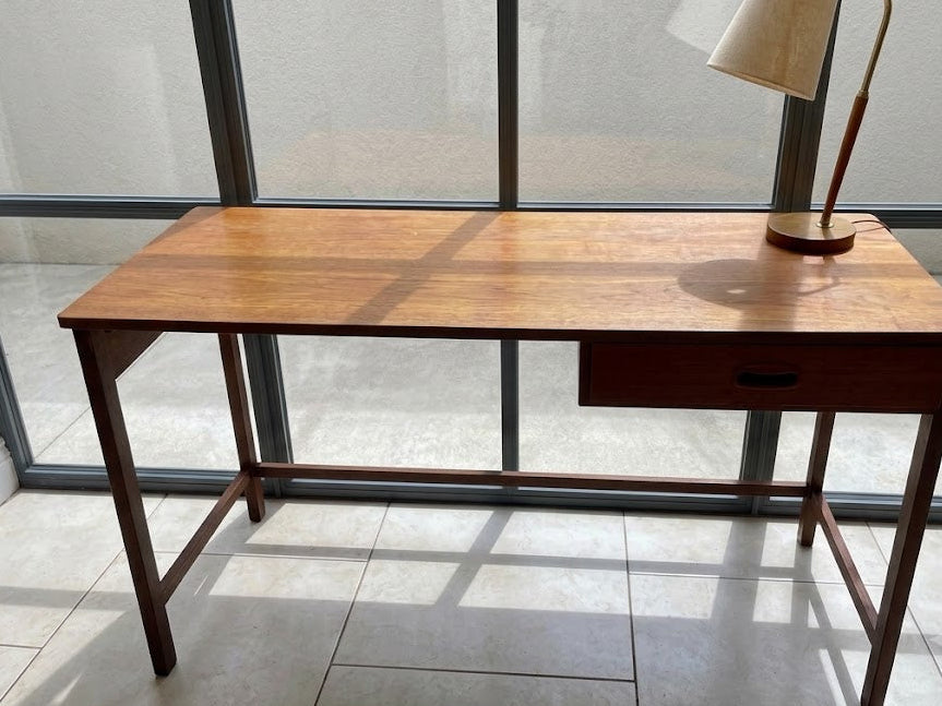 A mid-century modern wooden writing desk with a single drawer and a matching floor lamp on top, positioned in front of a large glass window.