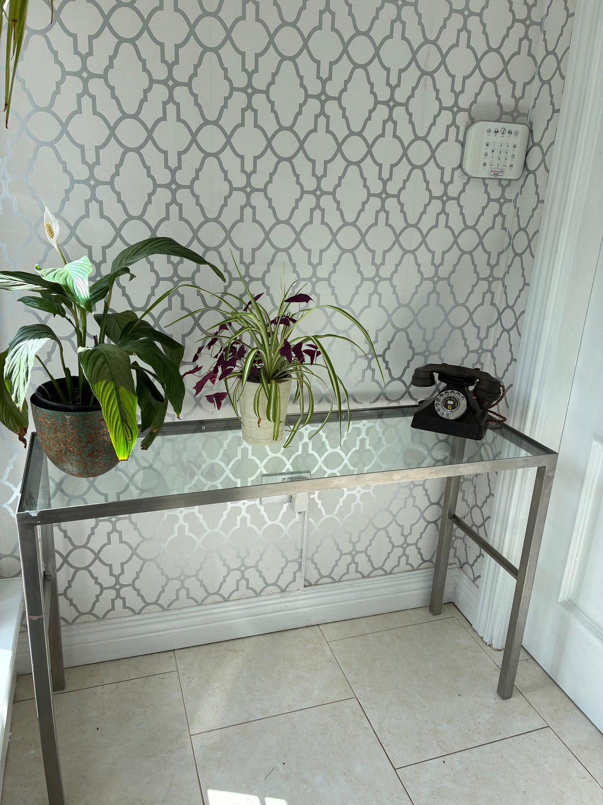 A modern glass-top console table with a silver metal frame, styled in a home entryway with two potted plants and a black rotary telephone against a white geometric patterned wallpaper background.
