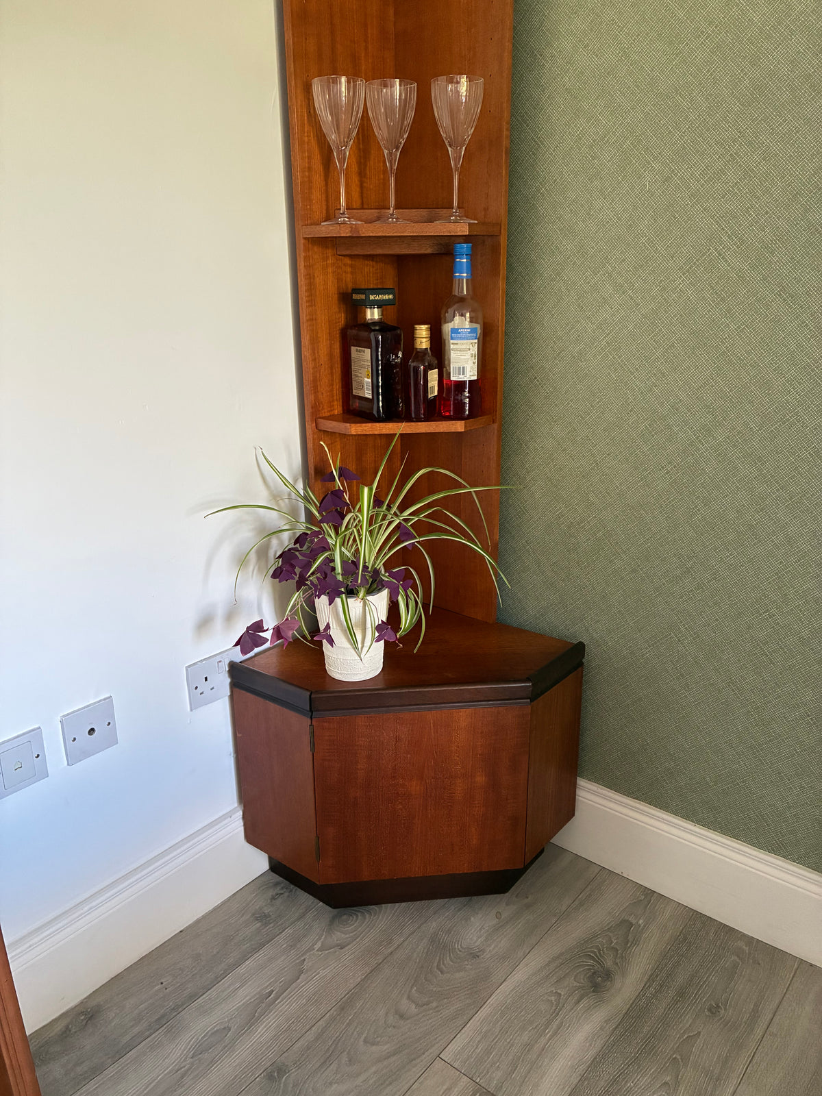 A corner wooden g plan cabinet with built-in shelves, styled with wine glasses, bottles, and a potted plant, set in a room with a green textured wall and gray wood-look floor.