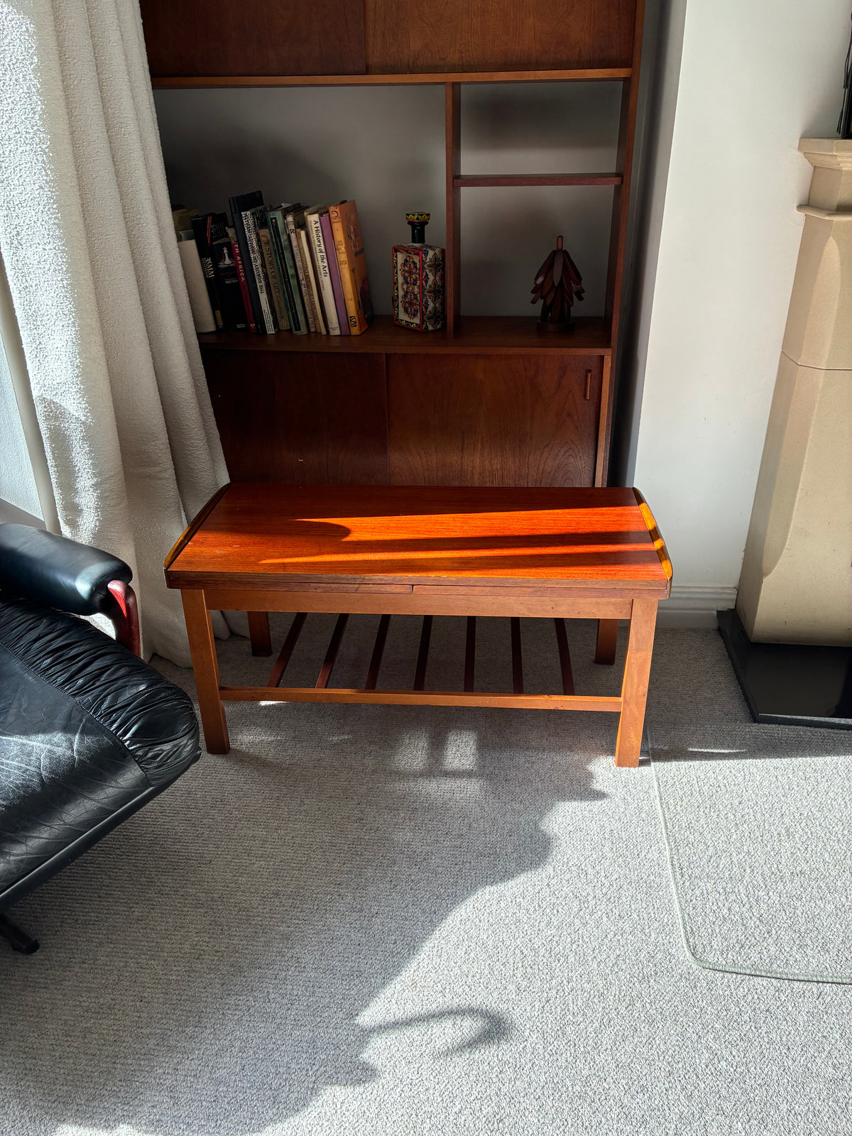 A mid-century modern wooden bench with a warm orange-brown finish and dark inlay stripes, positioned in a living room next to a black leather armchair and a wooden bookcase.