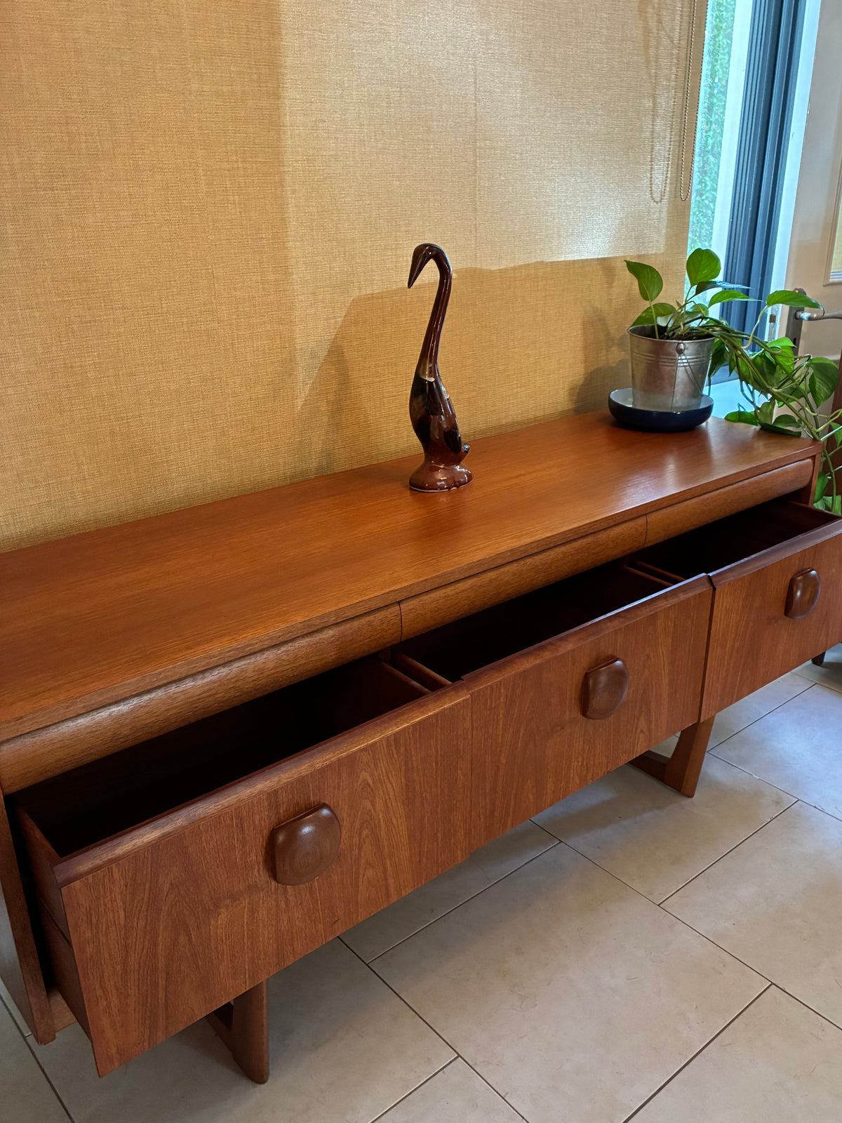 A mid-century modern wooden sideboard console with two open drawers, placed against a textured beige wall in a room with tiled floors. Decorative items including a dark metal bird sculpture and a potted plant sit on top.