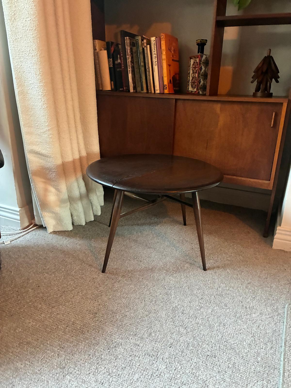 A mid-century modern ercol wooden side table with a round dark top and angled legs, positioned in a room with a beige carpet, a curtain, and a mid-century credenza in the background.
