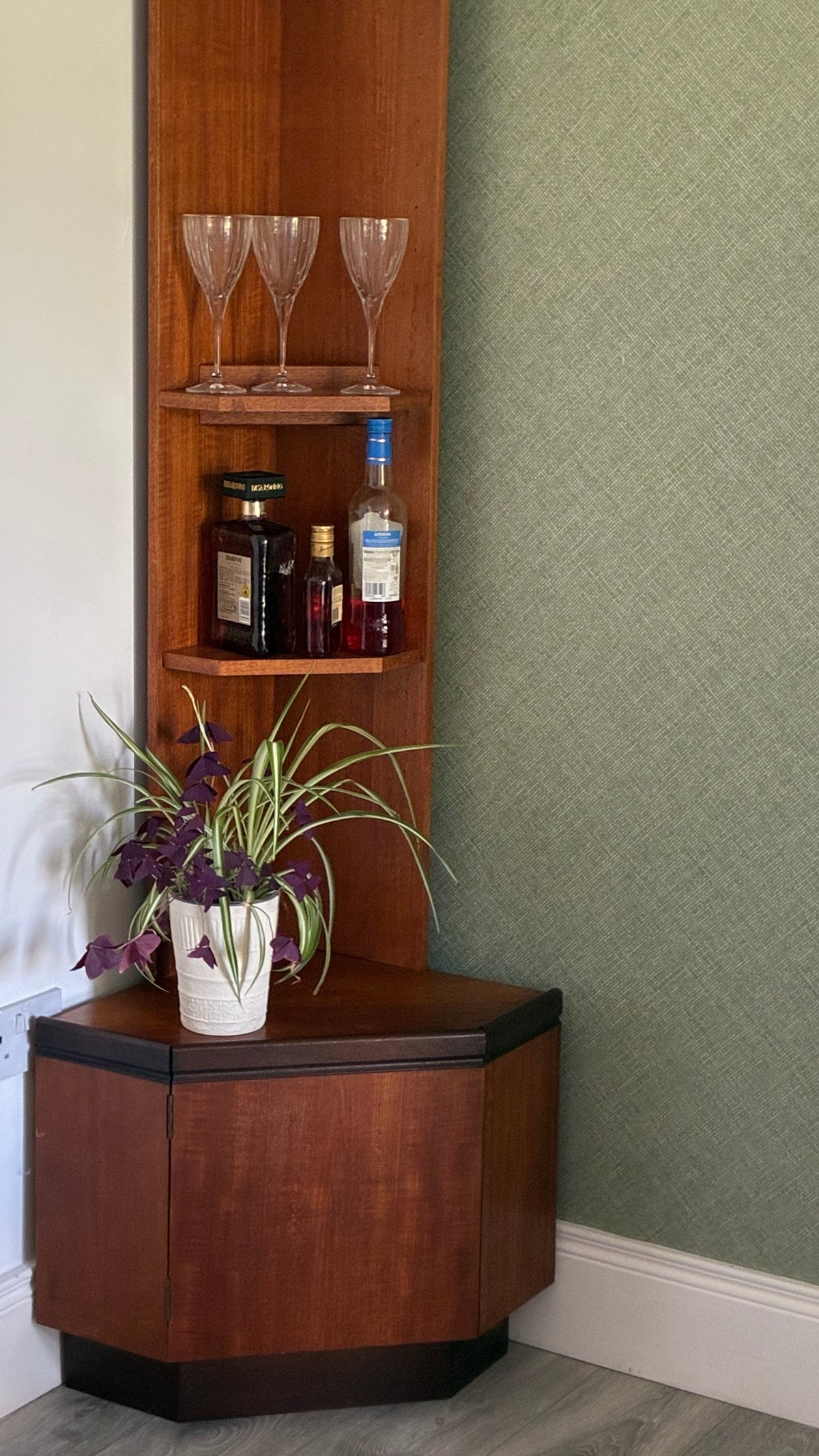 A tall, narrow g plan wooden hutch or shelf unit with three open shelves and a lower hexagonal cabinet, styled with wine glasses, bottles, and a potted plant in a room corner against a green textured wall.