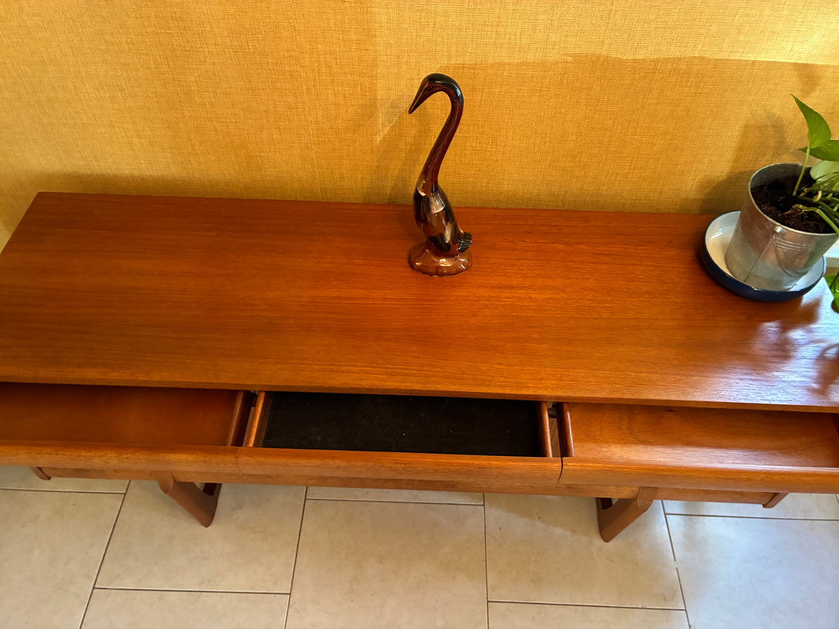 A mid-century modern wooden console table with a warm reddish-brown finish, featuring a swan figurine on top and a potted plant on the right side, set against a textured yellow wall and tiled floor.
