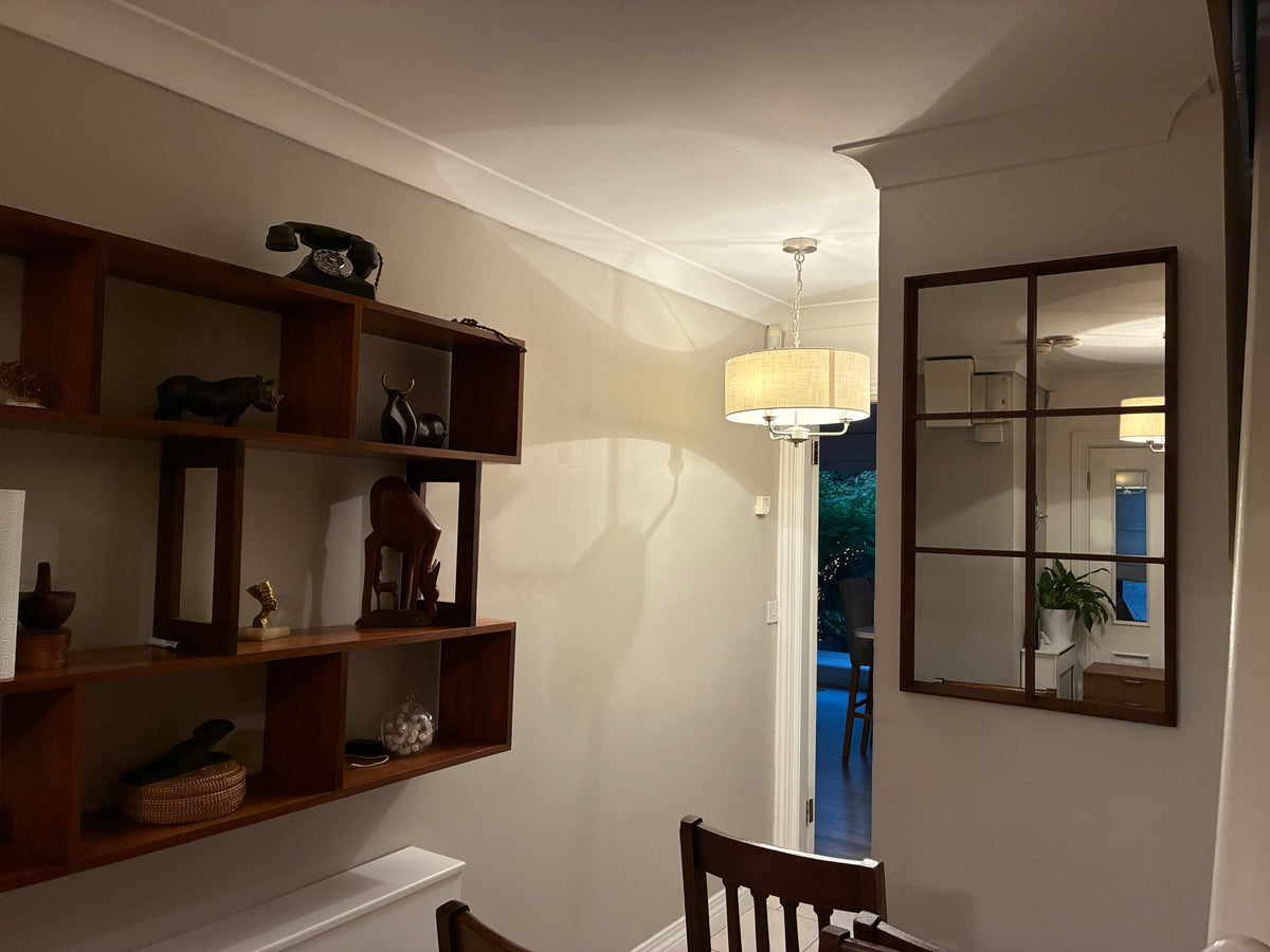 A warm, dimly lit interior corner featuring a dark brown floating wooden shelf on the left, a matching wall mirror with a dark wood frame on the right, and a hanging cream-colored drum lamp in the center.