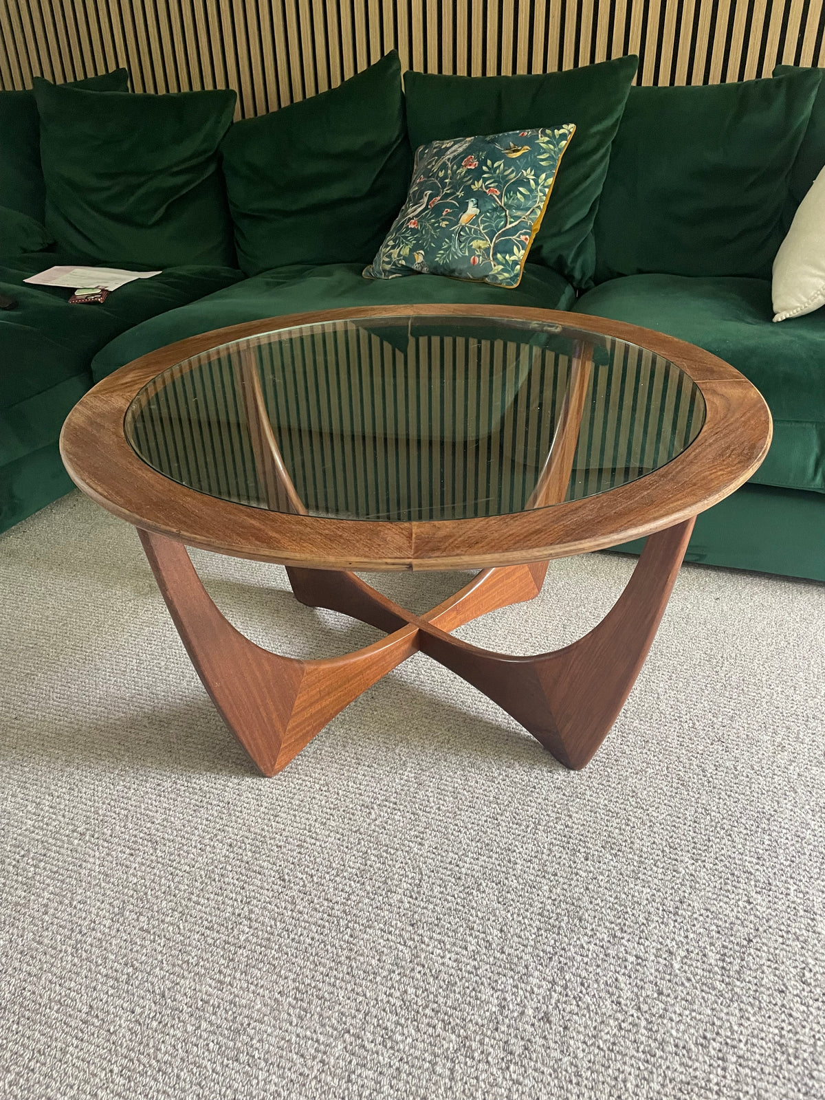A mid-century modern oval coffee table with a glass top and sculptural wooden legs, set in a living room in front of a green velvet sofa.