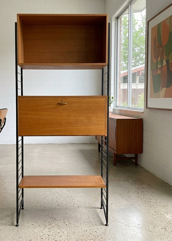 A mid-century modern wooden shelving unit with a three-tier open wood cabinet, a central drawer with a small metal pull, and a lower shelf, set against a white brick wall in an office environment.