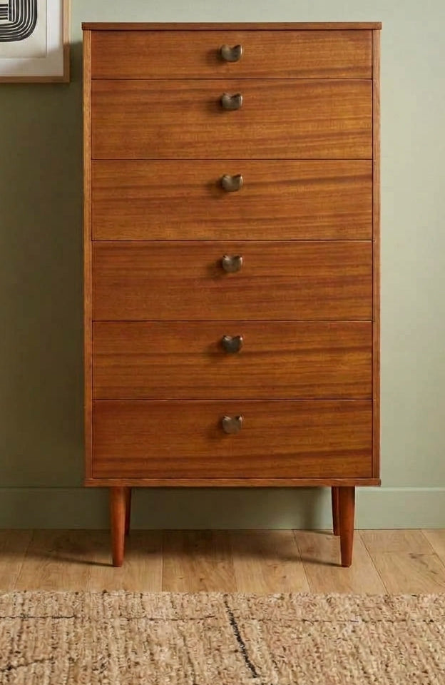 A mid-century modern wooden chest of drawers with five drawers and round metal knobs, set against a sage green wall with framed abstract art and a woven rug.