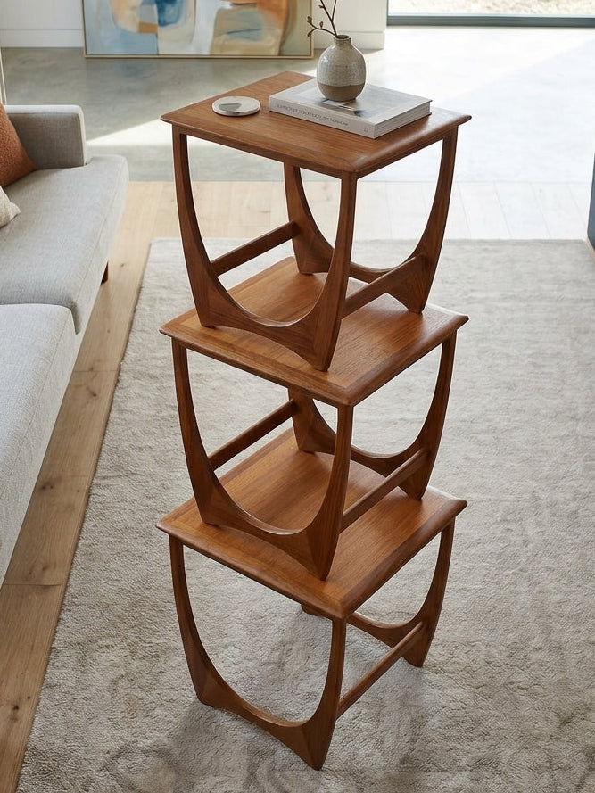 A mid-century modern wooden three-tier side table with curved sculptural legs, styled in a bright living room on a light rug next to a gray sofa.