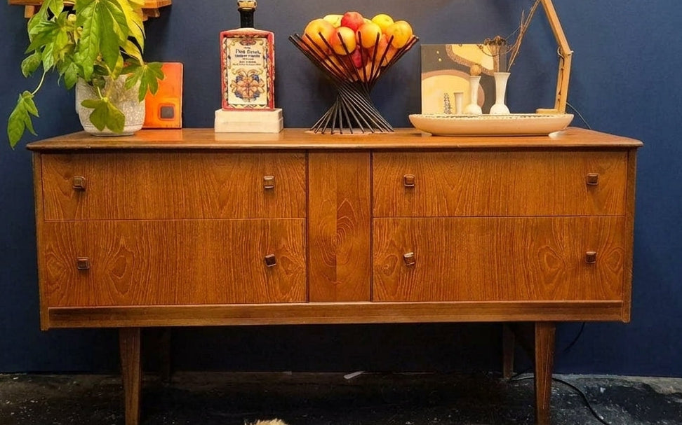 A mid-century modern wooden sideboard cabinet with six drawers, styled with books, a plant, and decorative objects against a dark blue wall.