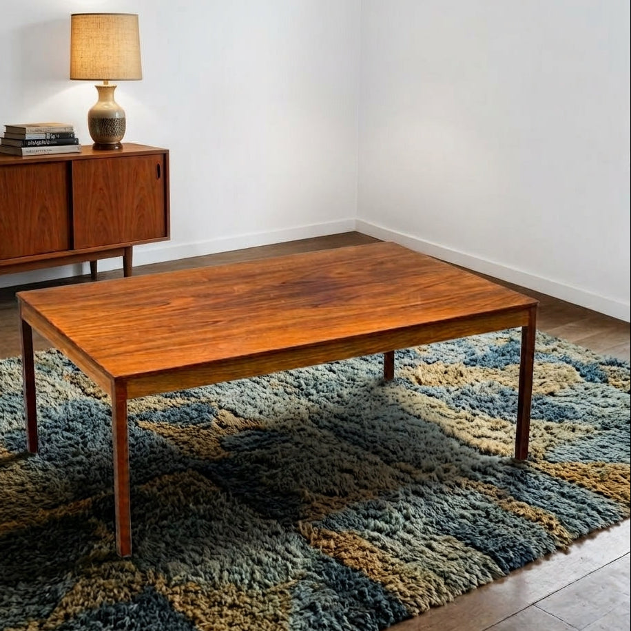 A rectangular mid-century modern wooden coffee table with a warm medium-brown finish, set on a textured blue and beige shag rug in a minimalist room with a mid-century sideboard and lamp in the background.