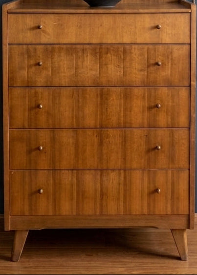 A mid-century modern wooden chest of drawers with five drawers, styled in a room with a dark blue wall, an oval mirror, a floor lamp, and a woven wall hanging.