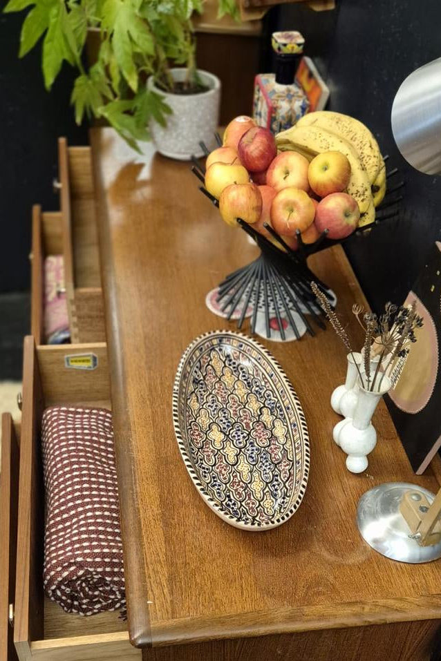 A wooden dresser or console table with open drawers, styled with a fruit bowl, a decorative oval tray, a small white vase, and a silver desk lamp in a home interior setting.