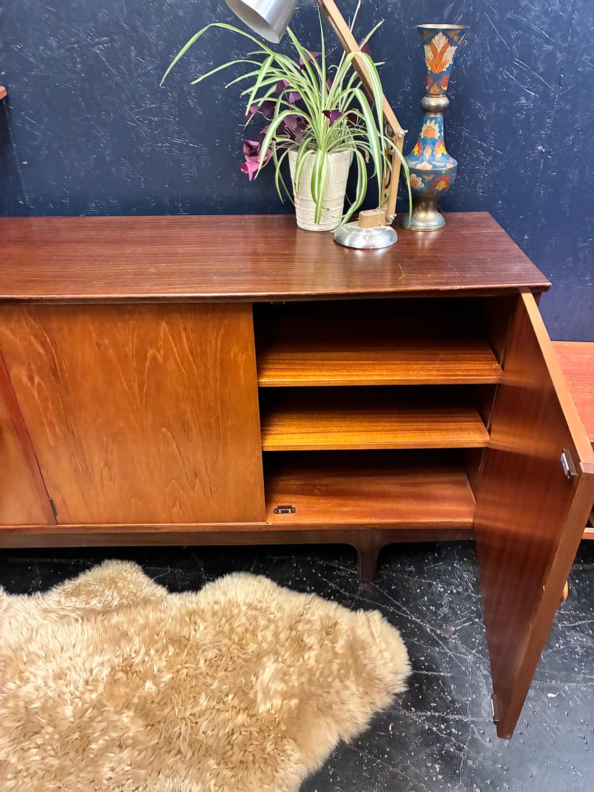 A mid-century modern wooden sideboard cabinet with a warm reddish-brown finish, shown with its right door open to reveal three interior shelves. The cabinet is styled with a potted plant, a metal lamp, and a decorative vase against a dark textured wall, with a shaggy beige rug on the floor.