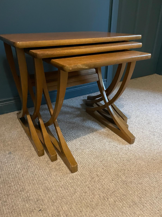 Three nested wooden nesting tables with a warm medium-brown finish, set on a beige carpet against a dark teal wall.