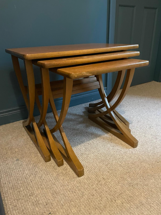 Three nested wooden nesting tables with a warm medium-brown finish, set on a beige carpet against a dark teal wall.
