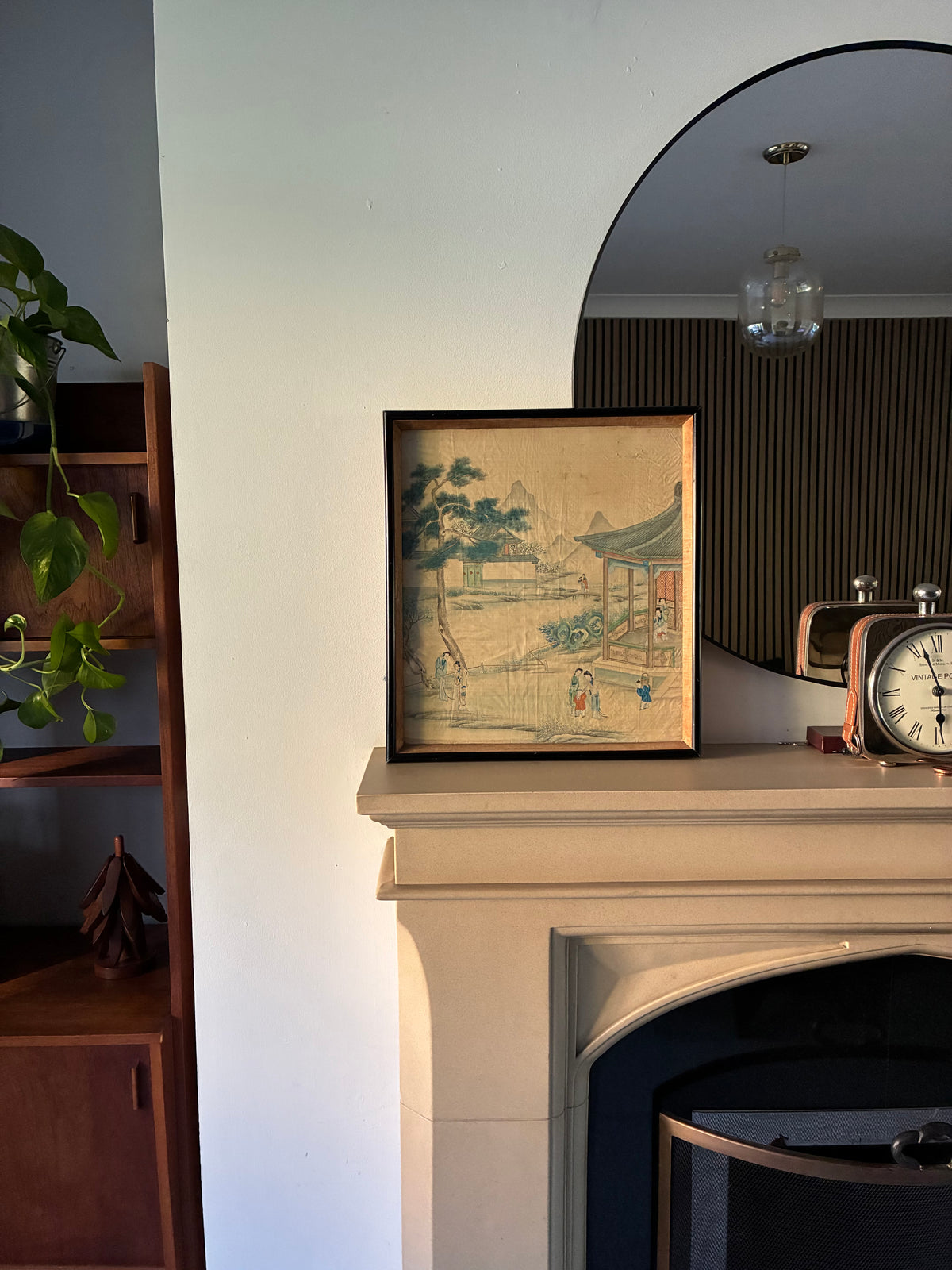 A framed vintage-style landscape art print with an Asian-inspired scene, displayed on a cream fireplace mantel in a home interior next to a wooden shelf and an arched mirror.