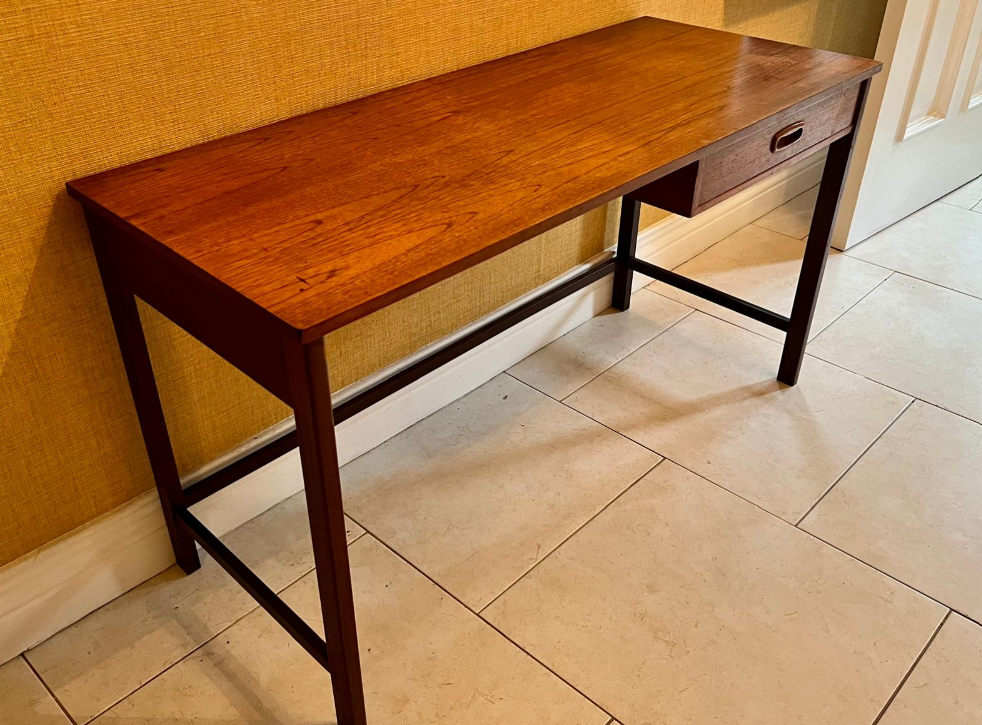 A mid-century modern wooden writing desk with a single drawer, positioned in a home setting against a textured yellow wall and tiled floor.