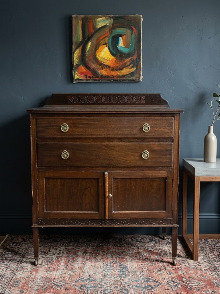 A dark brown wooden hutch or sideboard cabinet with two drawers and two lower doors, shown in a styled room setting against a dark blue wall with an abstract painting above and a patterned rug underneath.