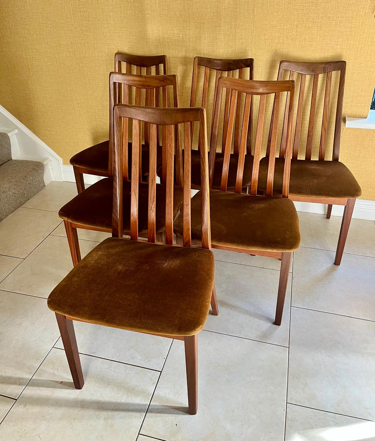 Six wooden dining chairs with brown velvet seats and backs, arranged on a light tiled floor against a textured yellow wall.