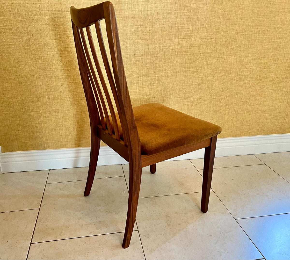 A mid-century modern wooden dining chair with a slatted backrest and a brown upholstered seat, shown in a room with a textured yellow wall and tiled floor.