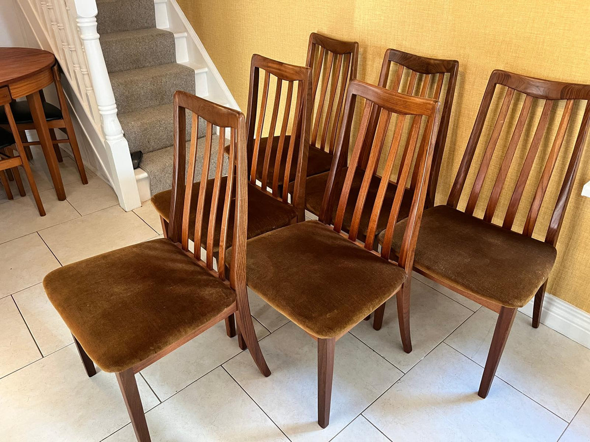 Six wooden dining chairs with brown upholstered seats and backs, arranged on a tiled floor in a home interior next to a staircase.