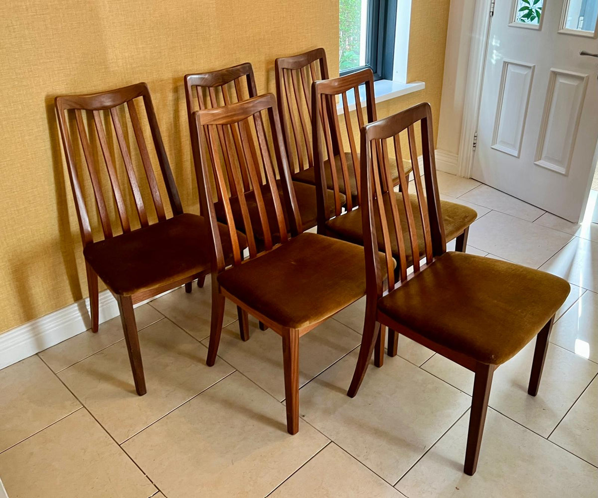 Six wooden dining chairs with slatted backs and brown upholstered seats, arranged on a tiled floor against a yellow wall.