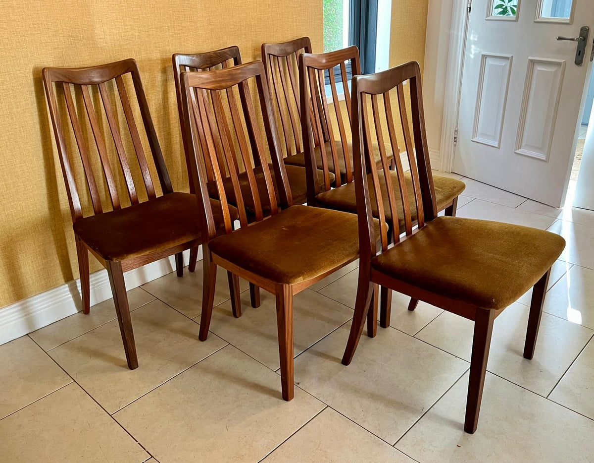 6 mid-century G Plan modern wooden dining chairs with vertical slat backs and brown velvet seats, arranged on a tiled floor against a textured yellow wall.