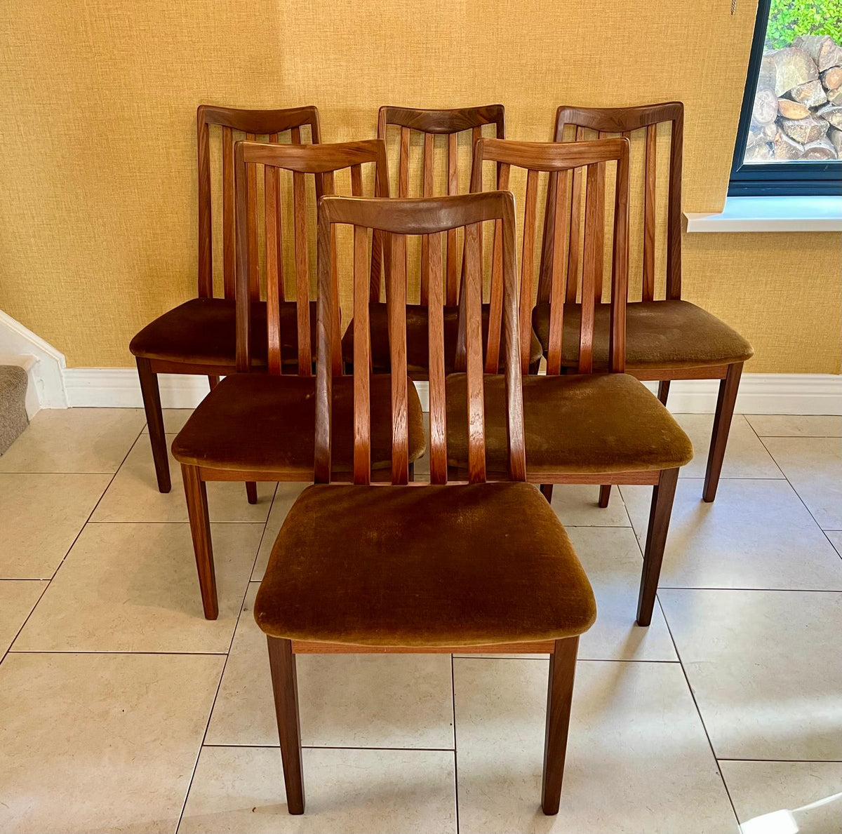 Six wooden dining chairs with brown upholstered seats arranged in a group on a tiled floor against a textured yellow wall.