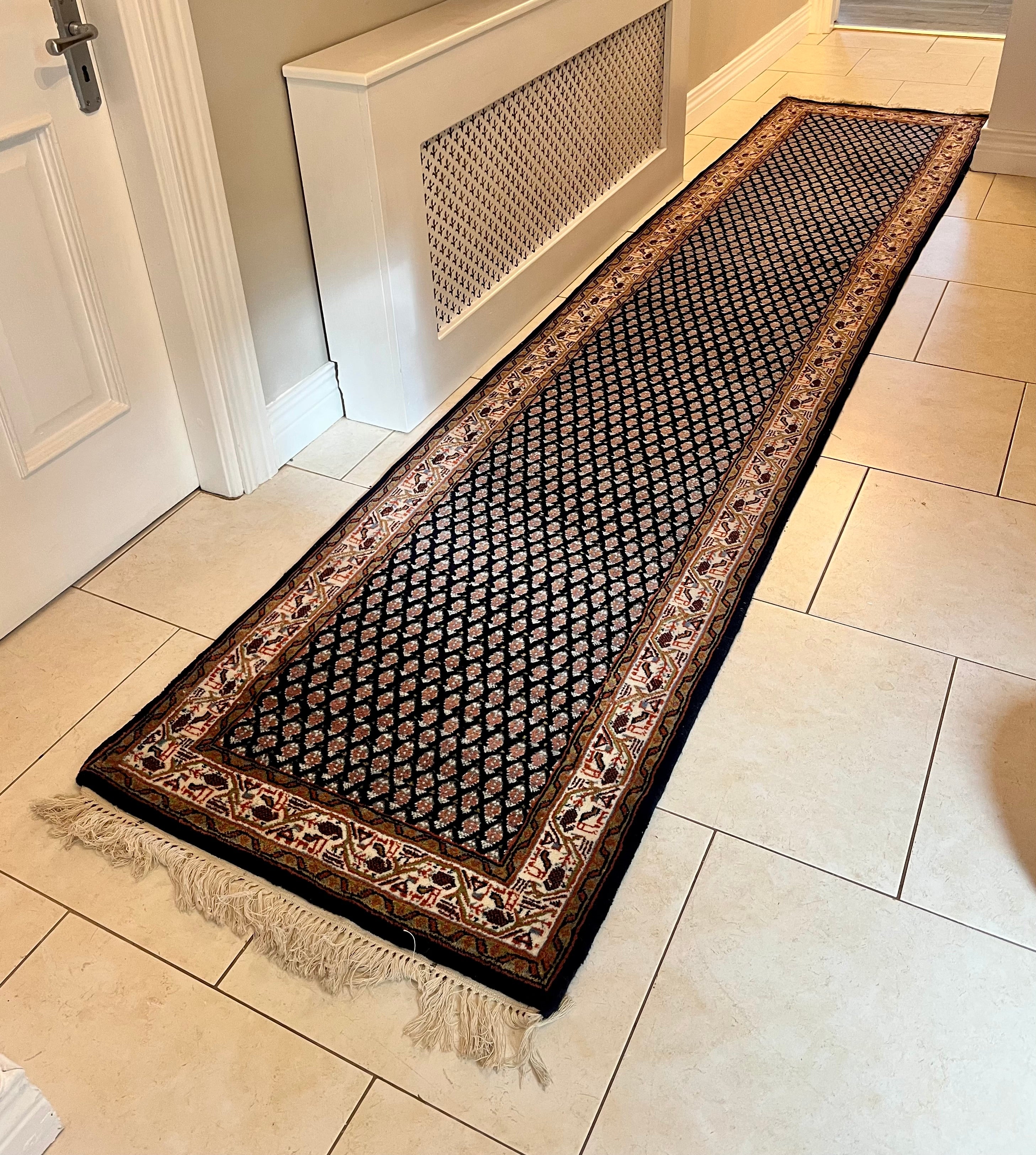 A handmade vintage patterned area rug with a dark navy blue field, cream and orange geometric motifs, and a detailed border, shown laid out on a light-coloured tiled floor with fringe visible at the bottom by Grace & Joy Furniture