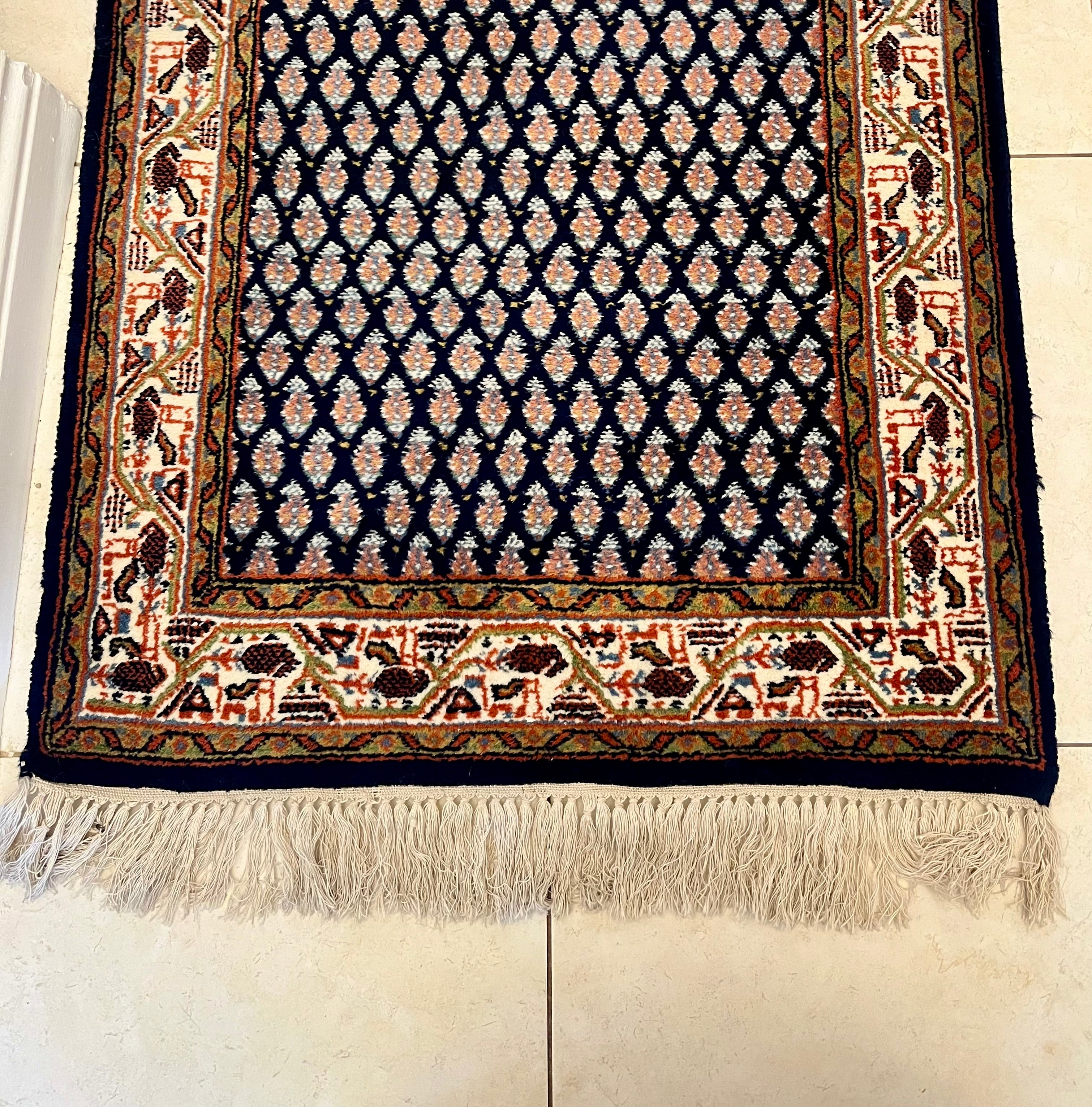 A handmade vintage patterned area rug with a dark navy blue field, cream and orange geometric motifs, and a detailed border, shown laid out on a light-coloured tiled floor with fringe visible at the bottom by Grace & Joy Furniture