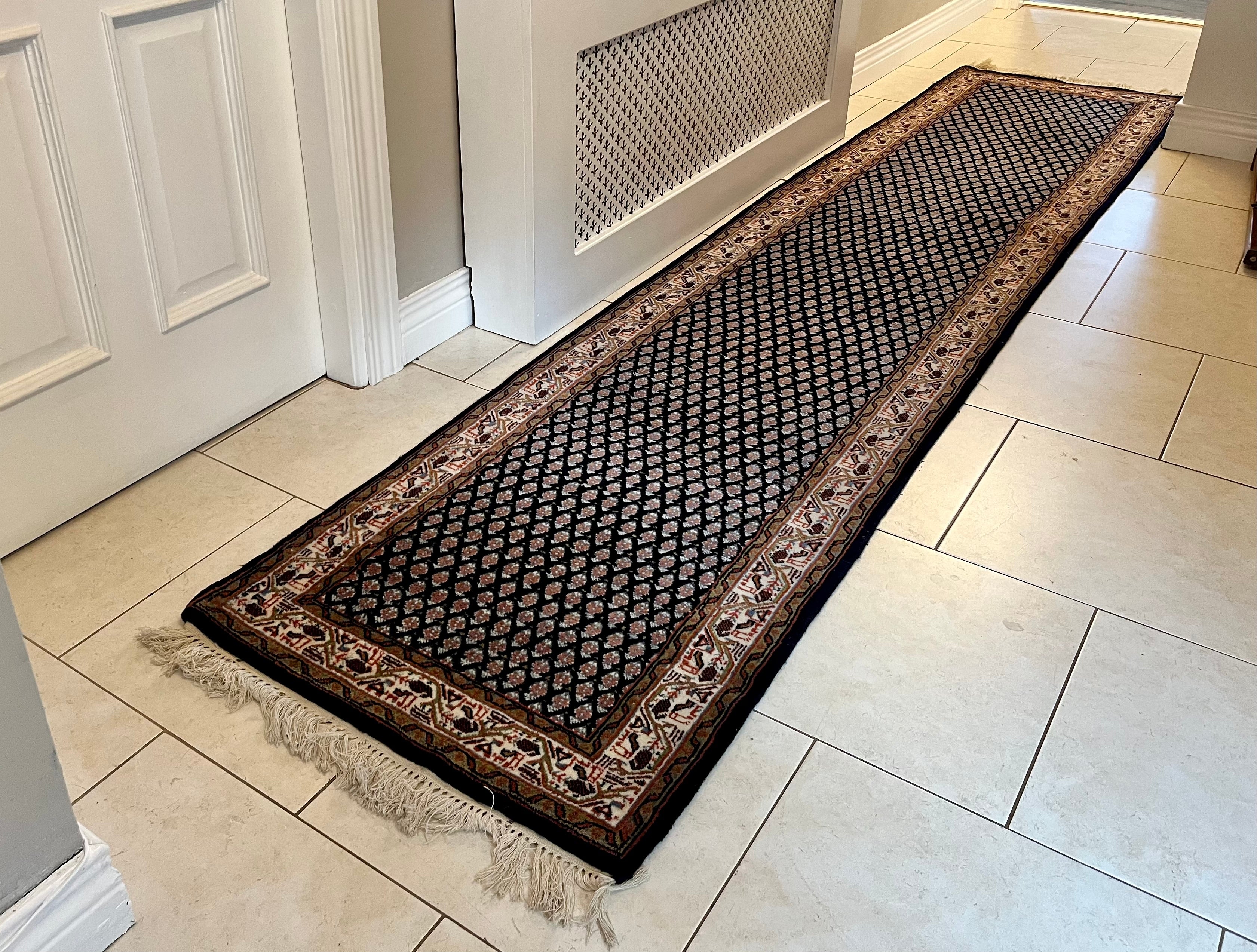 A handmade vintage patterned area rug with a dark navy blue field, cream and orange geometric motifs, and a detailed border, shown laid out on a light-coloured tiled floor with fringe visible at the bottom by Grace & Joy Furniture