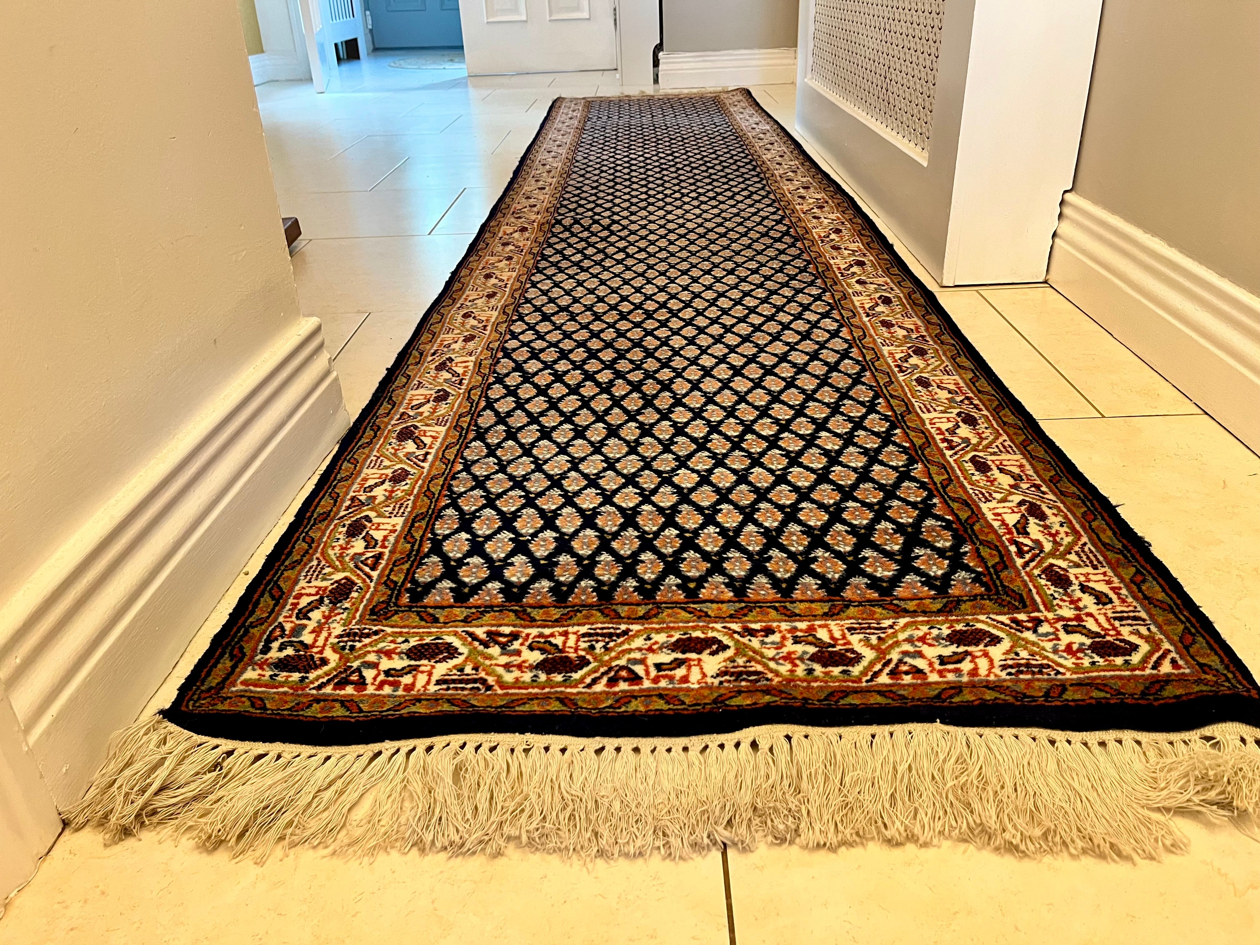 A handmade vintage patterned area rug with a dark navy blue field, cream and orange geometric motifs, and a detailed border, shown laid out on a light-coloured tiled floor with fringe visible at the bottom by Grace & Joy Furniture