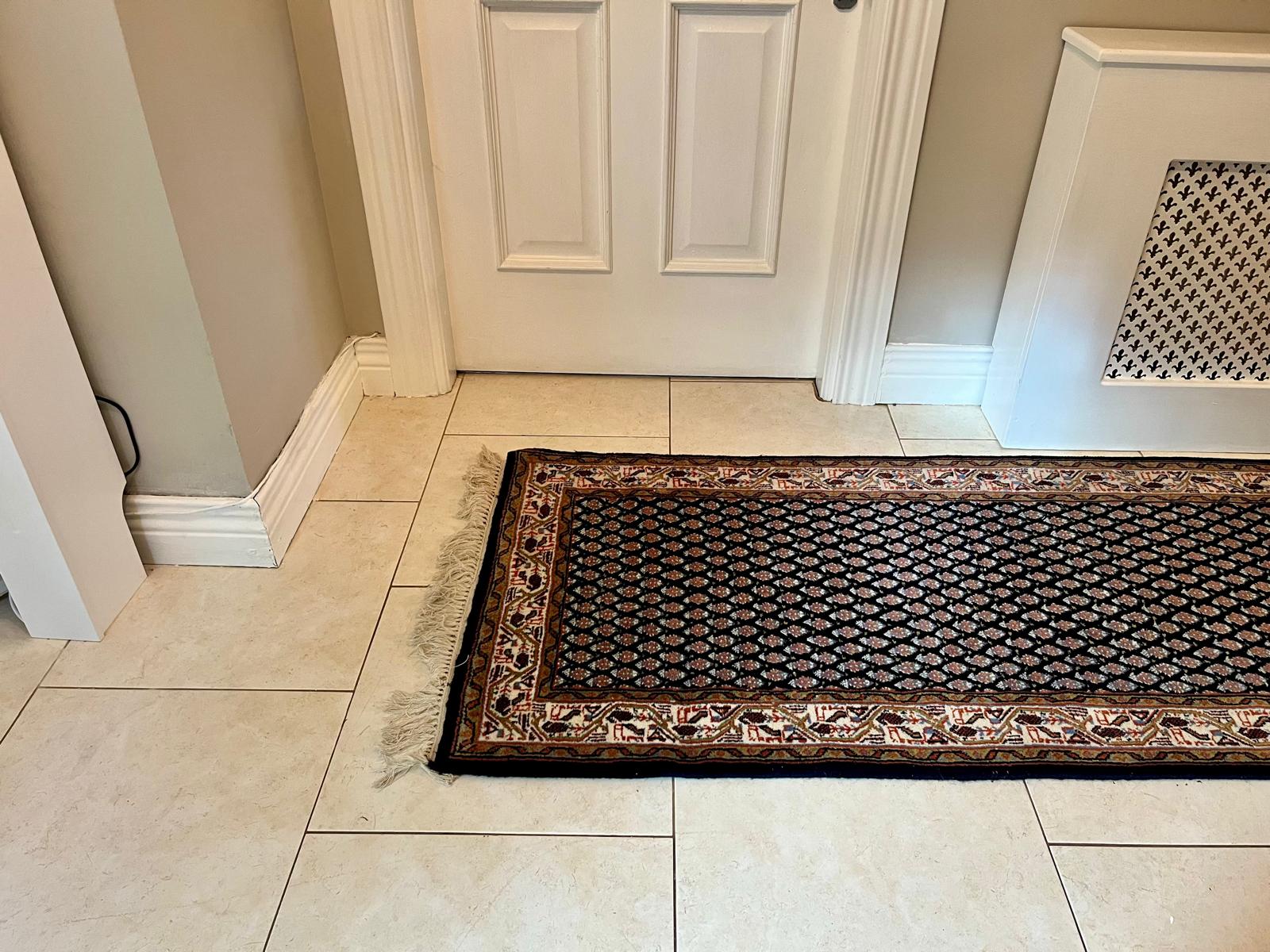 A handmade vintage patterned area rug with a dark navy blue field, cream and orange geometric motifs, and a detailed border, shown laid out on a light-coloured tiled floor with fringe visible at the bottom by Grace & Joy Furniture