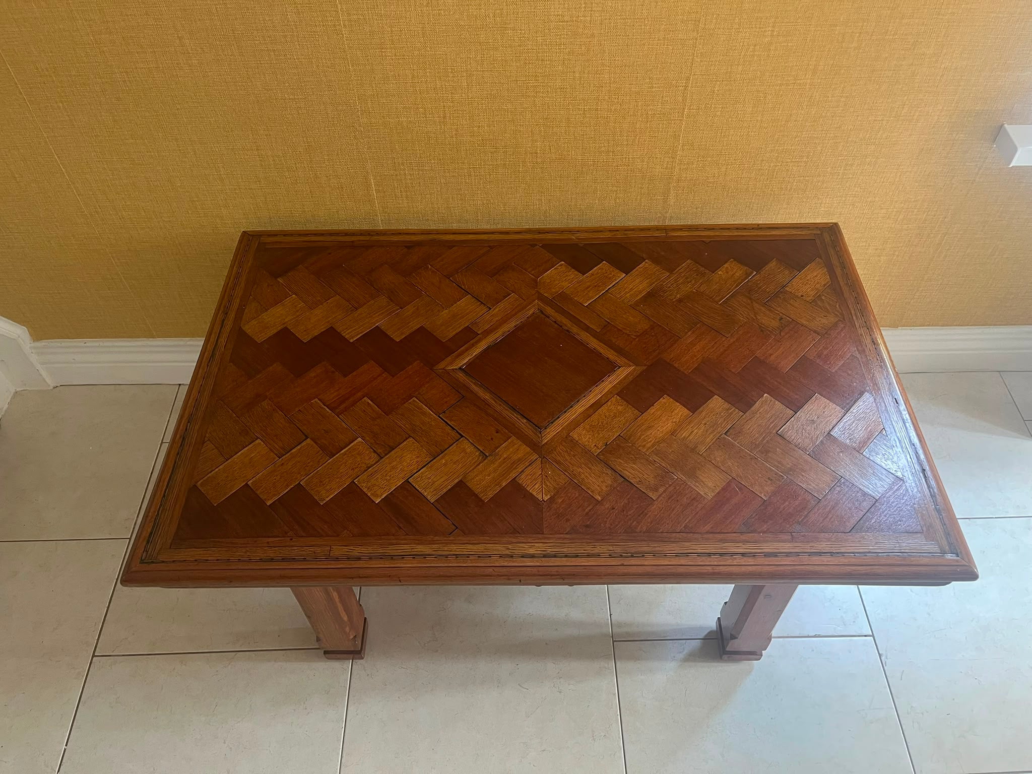 A Herringbone wooden coffee table with a geometric parquet inlay design on the top, set against a textured yellow wall and light-colored tiled floor by Grace & Joy Furniture
