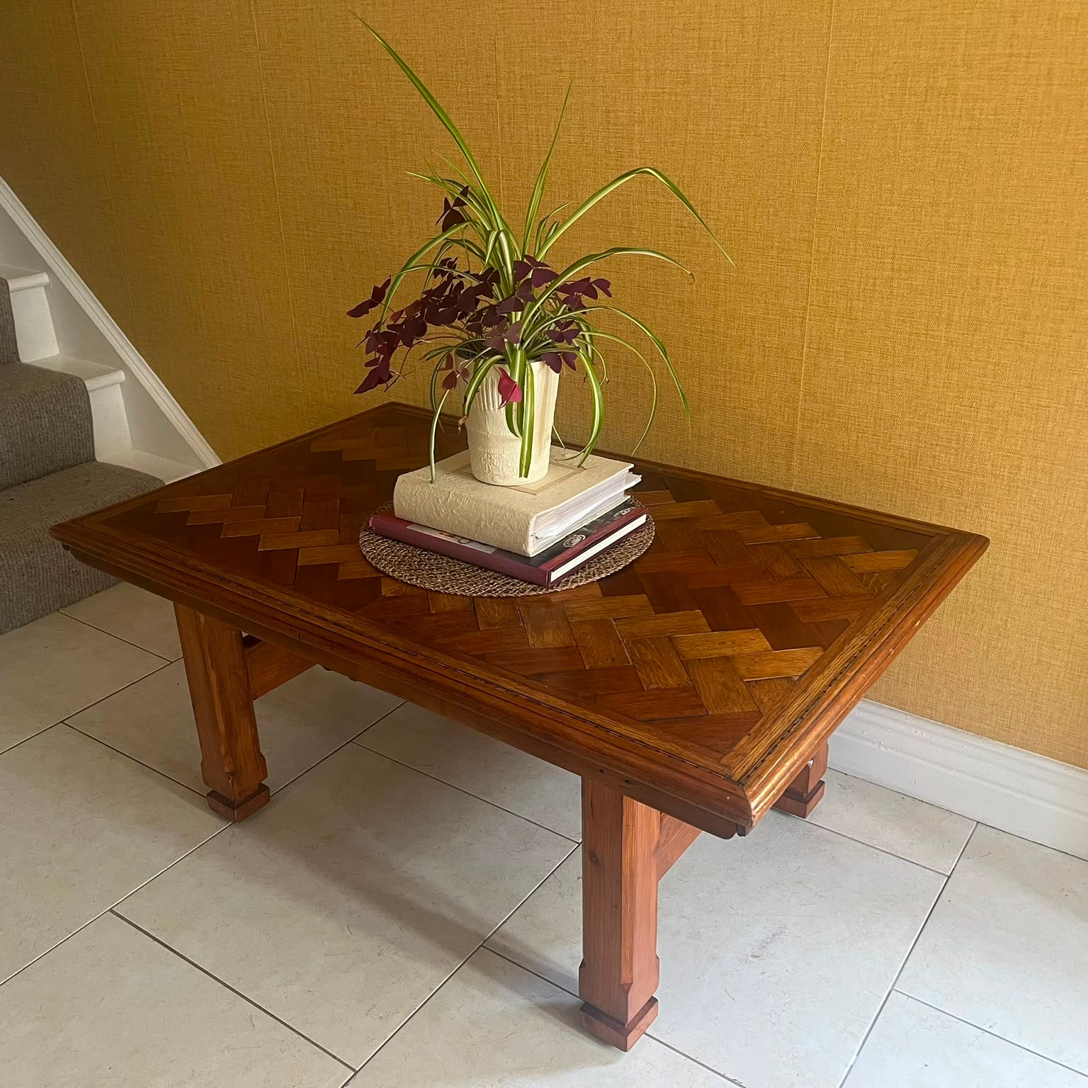 A Herringbone wooden coffee table with a geometric parquet inlay design on the top, set against a textured yellow wall and light-colored tiled floor by Grace & Joy Furniture