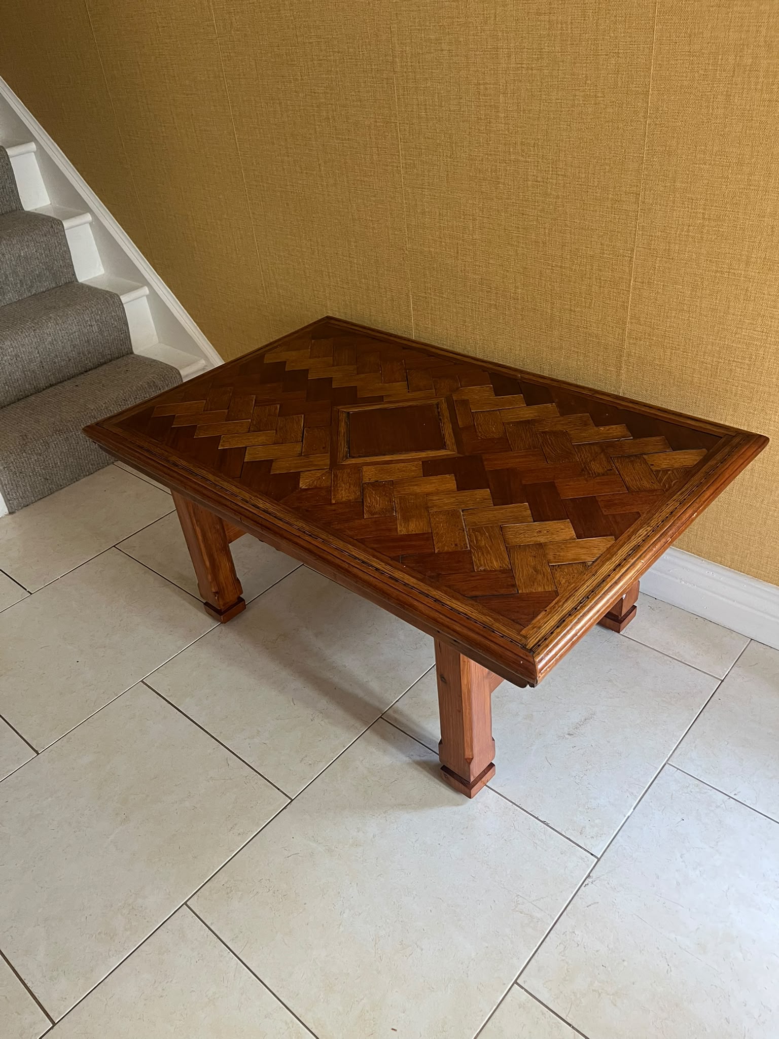 A Herringbone wooden coffee table with a geometric parquet inlay design on the top, set against a textured yellow wall and light-colored tiled floor by Grace & Joy Furniture