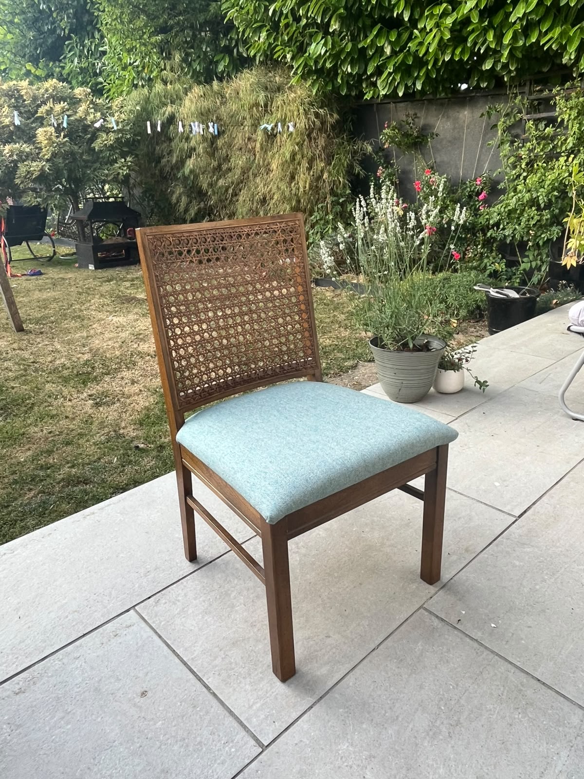 A wooden dining chair with a woven cane backrest and a light blue-green upholstered seat cushion, placed on a concrete patio in a garden setting.