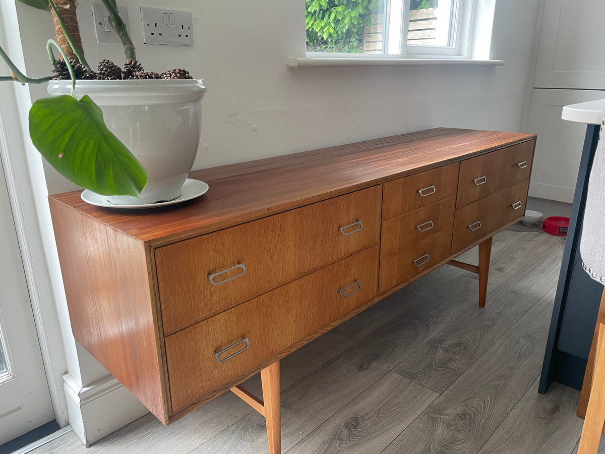Mid-century modern wooden meredew credenza with six drawers and silver handles, shown in a styled room with gray wood floors, a green wall, and a mirror behind it.