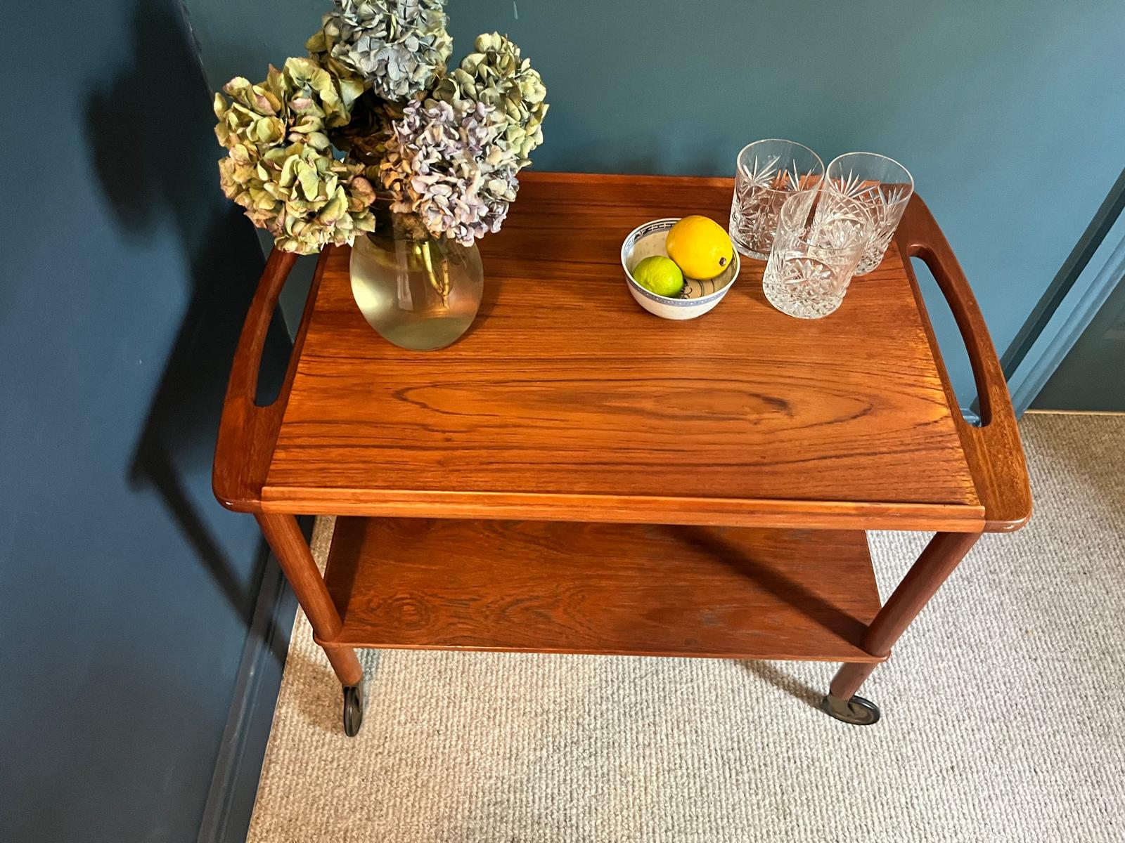 A mid-century teak Bowa drinks trolley, styled with a glass vase of hydrangeas, a small bowl of lemons, and two cut-crystal glasses against a dark teal wall and beige carpet by Grace & Joy Furniture