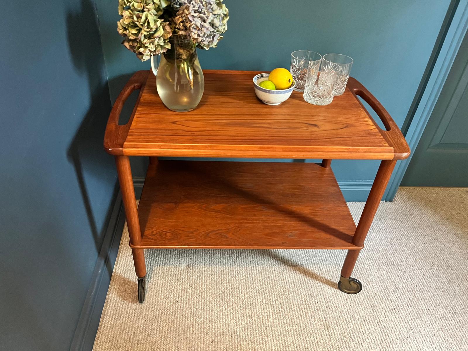 A mid-century teak Bowa drinks trolley, styled with a glass vase of hydrangeas, a small bowl of lemons, and two cut-crystal glasses against a dark teal wall and beige carpet by Grace & Joy Furniture