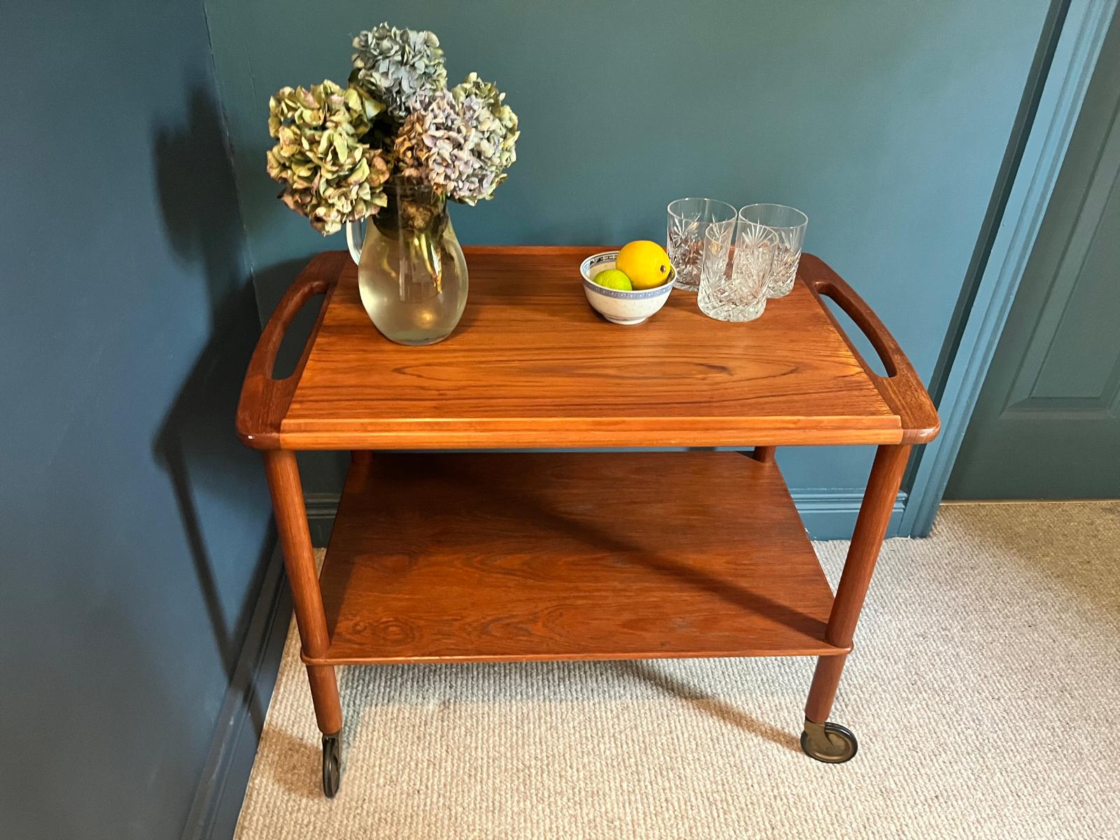 A mid-century teak Bowa drinks trolley, styled with a glass vase of hydrangeas, a small bowl of lemons, and two cut-crystal glasses against a dark teal wall and beige carpet by Grace & Joy Furniture