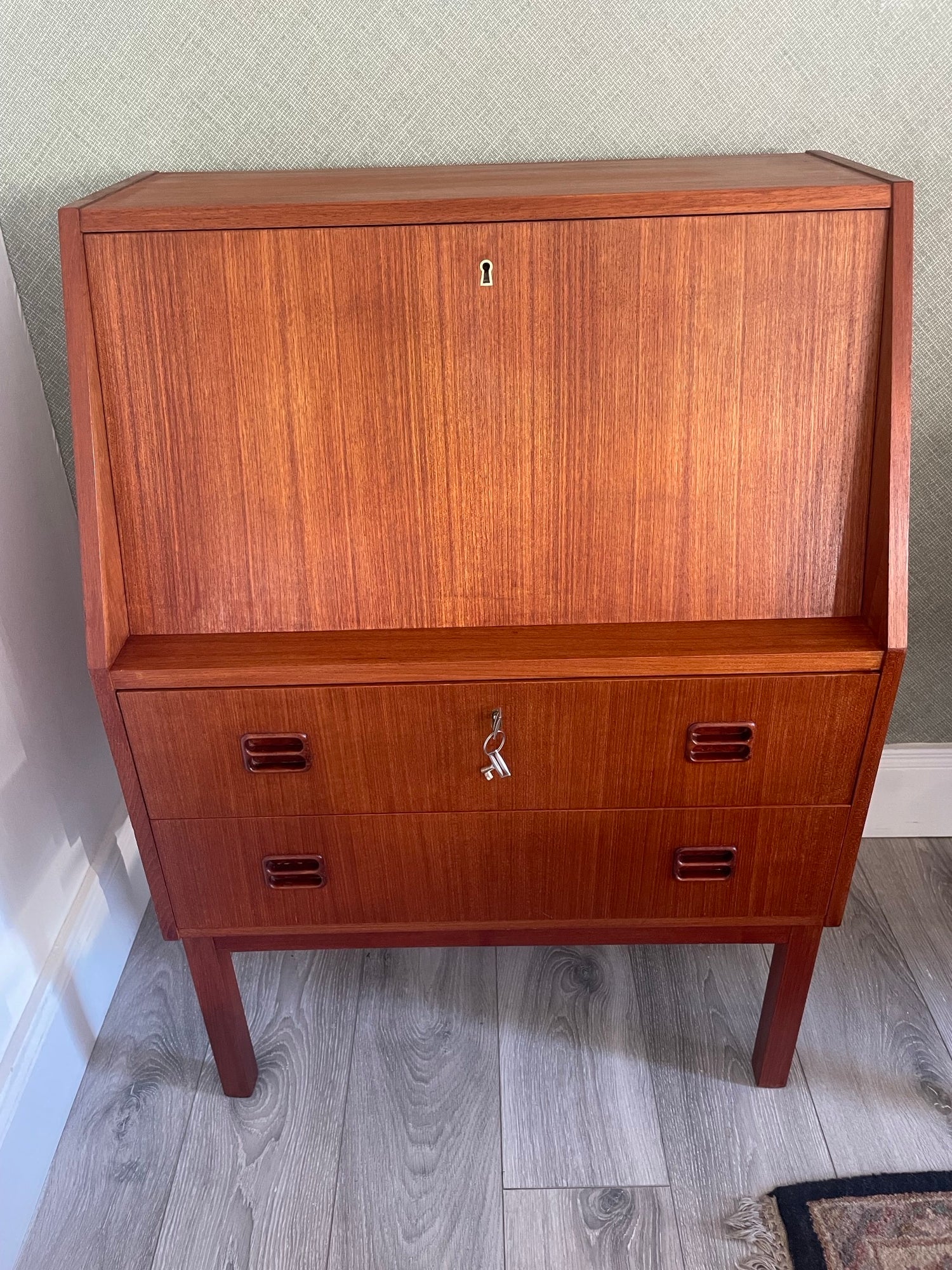 A mid-century modern Danish bureau with a warm reddish-brown finish, featuring a large left door and three drawers on the right with dark metal pulls, photographed indoors on a gray wood floor. by Grace & Joy Furniture