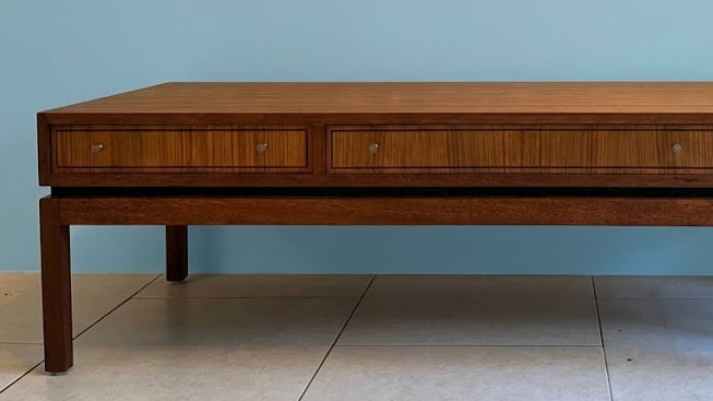 A mid-century modern wooden coffee table by Greaves & Thomas with three drawers and brass knobs, styled in a minimalist living room with a blue wall, a brown leather lounge chair, and a large window overlooking a garden by Grace & Joy Furniture